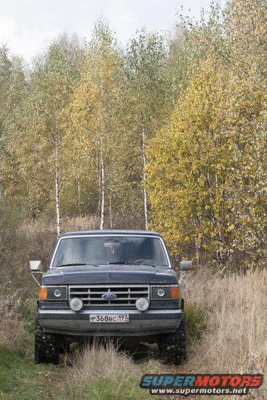 Dark blue 1980s Ford Bronco with off-road tires and auxiliary lights parked in a grassy area with autumn trees