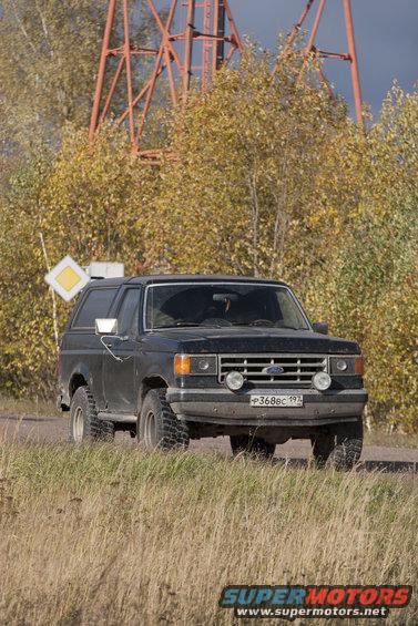 Black 1987 Ford Bronco parked on a dirt road with trees and a large metal structure in the background
