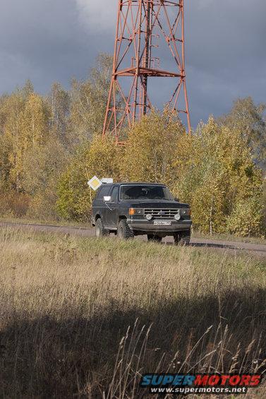 Dark gray Ford Bronco driving on a dirt road near a tall radio tower. Autumn trees in background