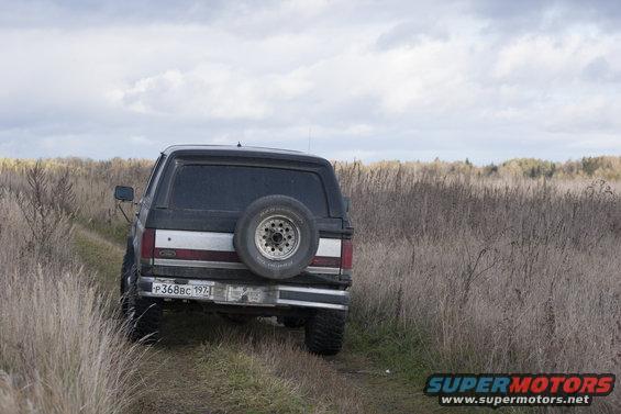 1987 Ford Bronco rear view, driving on a dirt path in a field of dry grass