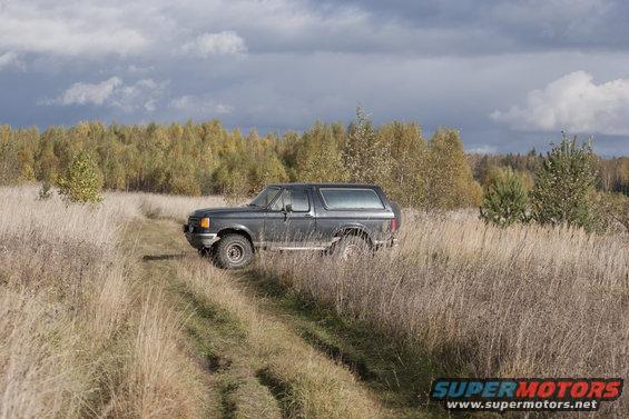 Dark gray 1987 Ford Bronco on a dirt path through tall dry grass, with autumn trees in the background