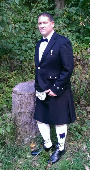 Man in a kilt and suit jacket stands next to a tree stump outdoors