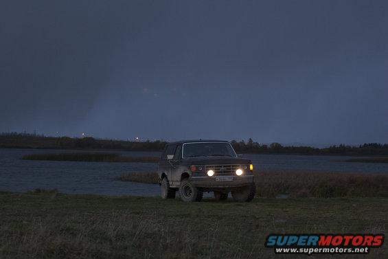 1987 Ford Bronco driving on a dirt road at dusk, headlights on