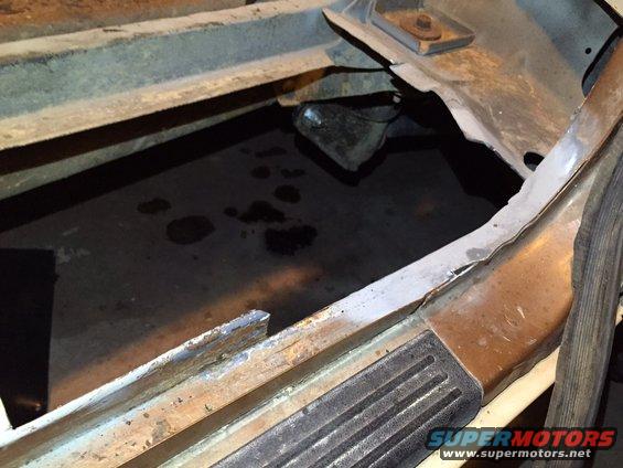 1991 Ford Bronco floor pan with significant rust and holes, showing a dark, wet surface below