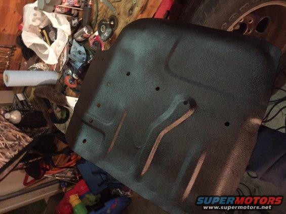 Black vehicle floor pan with some holes and raised ridges. Background is cluttered with tools and supplies