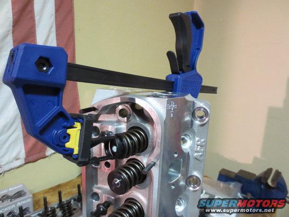 Engine cylinder head held by a clamp, showing valve springs and ports. Text visible on the part