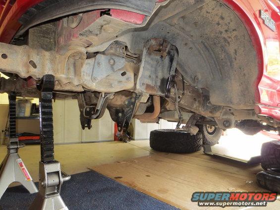 Red vehicle frame and suspension on jack stands, showing rust and undercarriage components