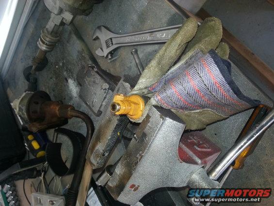 Yellow fuel injector clamped in a vise with a wrench and gloves nearby
