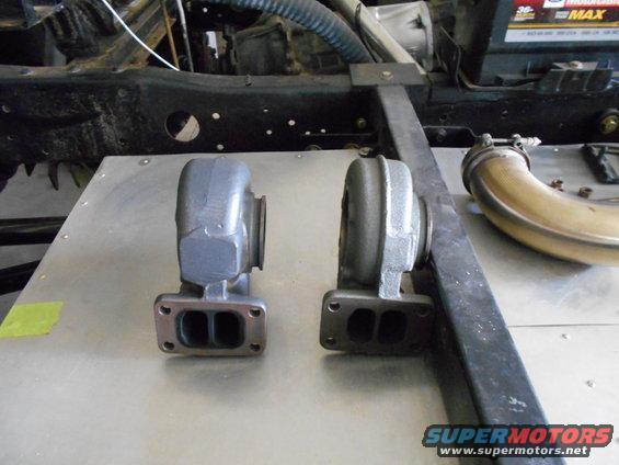 Two turbochargers and exhaust piping on a metal surface with vehicle frame in background