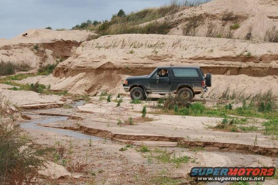 Dark 1987 Ford Bronco driving through a sandy, eroded landscape with a small stream