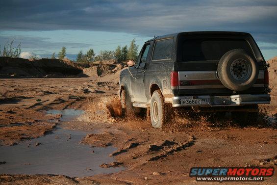 Black 1987 Ford Bronco driving through mud, rear view with spare tire mounted