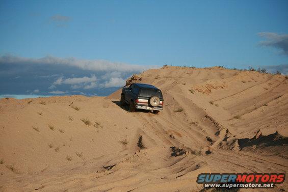 Black SUV with spare tire on back driving up a sand dune under a cloudy blue sky