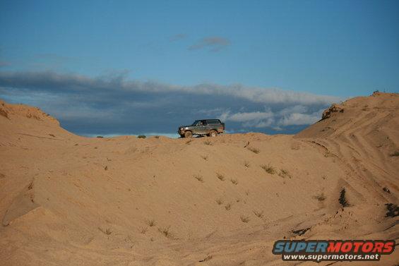 1987 Ford Bronco on a sand dune under a cloudy blue sky