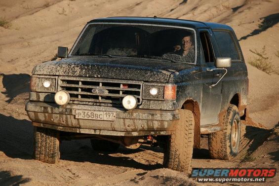 1987 Ford Bronco driving on a sandy, dirt road, covered in mud
