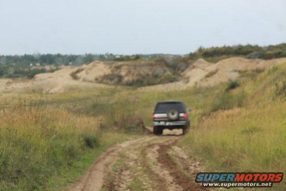 SUV with spare tire on rear door driving on muddy dirt road, grassy hills in background. SUPERMOTORS.NET logo