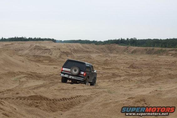 Black 1987 Ford Bronco driving on a sandy terrain with tire tracks visible