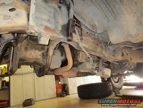 Underside view of vehicle frame, showing exhaust pipe, suspension parts, and a spare tire. The vehicle appears to be on a lift in a garage