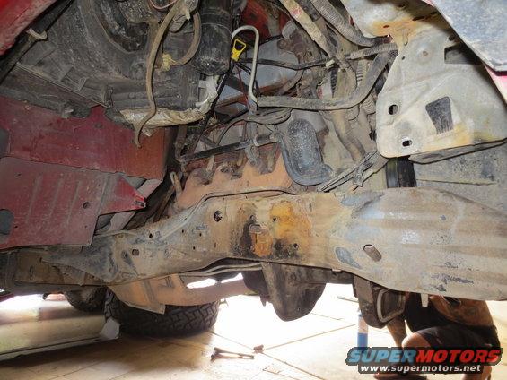 Underside view of a vehicle's engine and frame, showing rust and various mechanical parts