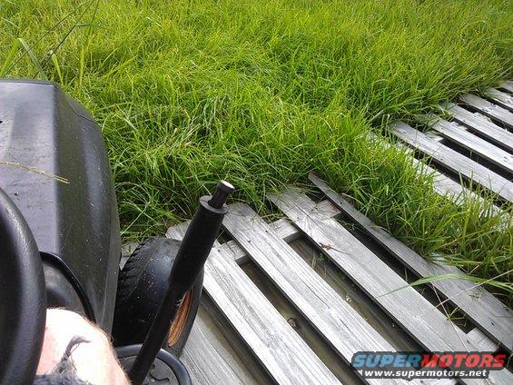 Lawn mower interior with black seat and control lever, next to tall grass and wooden planks