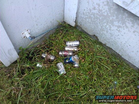 Aluminum cans scattered on grass near a white wall and a spigot