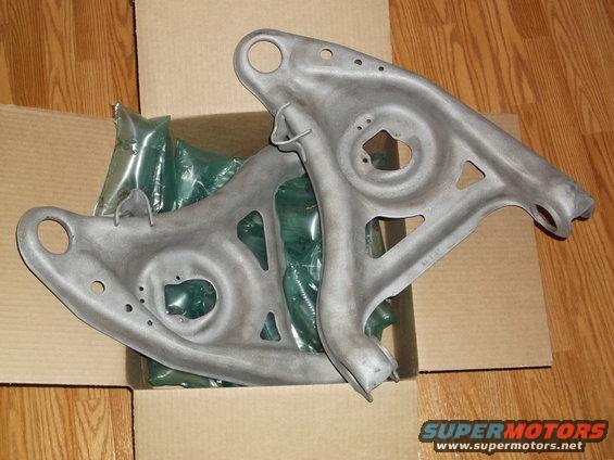 Two gray metal suspension control arms, one on top of the other, in a cardboard box with green packaging