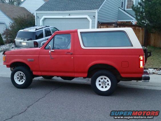 1988 Ford Bronco parked on street, side view. Silver SUV in driveway behind it