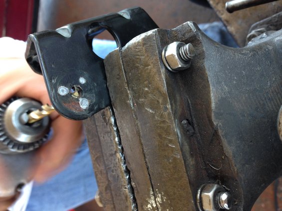 Drill bit entering a black metal bracket secured in a vise. The bracket has three small white marks near the hole
