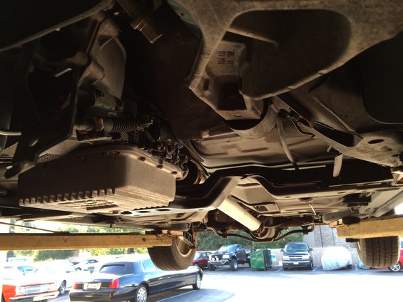 Underside view of a car on a lift, showing the transmission and exhaust. Other cars are parked in the background