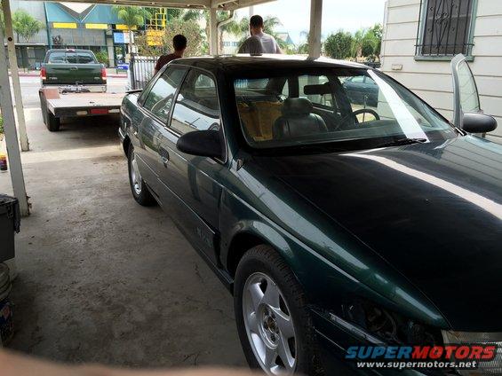 Dark green sedan parked under a carport, with a green pickup truck and trailer behind it