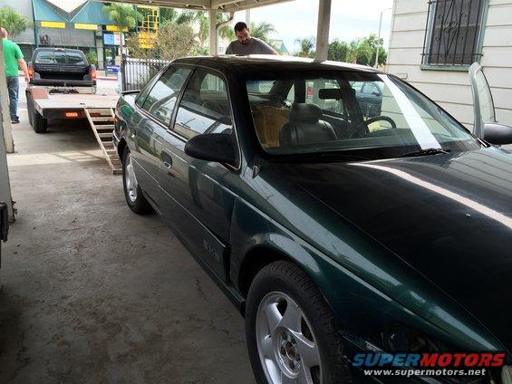 Dark green sedan parked under a carport, with a pickup truck and trailer in the background