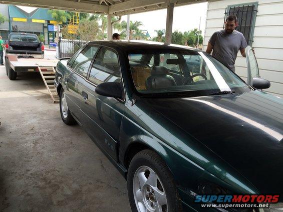 Green sedan with a man opening the driver's door, parked under a carport next to a trailer with a black pickup truck