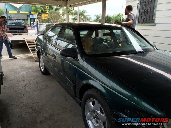 Dark green sedan parked under a carport, with a man standing by the driver's side window. Another man is by a trailer with a black truck