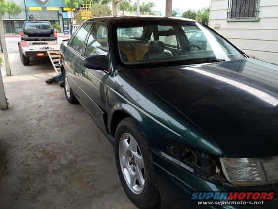 Dark green sedan parked next to a trailer with a black pickup on it. Headlight is missing