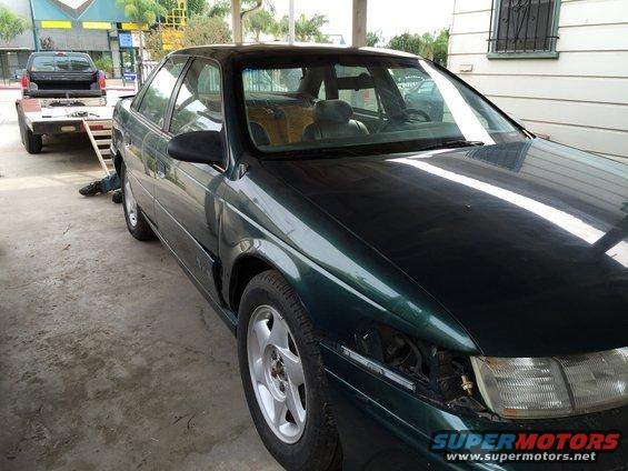 Green sedan with missing headlight, parked next to a trailer with a black pickup truck
