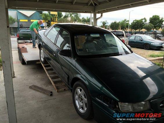 1993 Ford Taurus on a trailer with a man in a green shirt loading it; another car is parked in the background