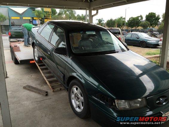 Dark green sedan being loaded onto a trailer. Another sedan is parked in the background