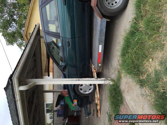 Green sedan being loaded onto a trailer under a carport. Man in green shirt is near the front wheel