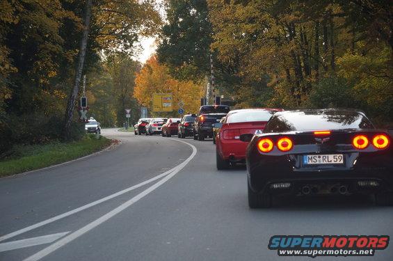 Several cars driving on a road with autumn trees. The closest car is black with red taillights