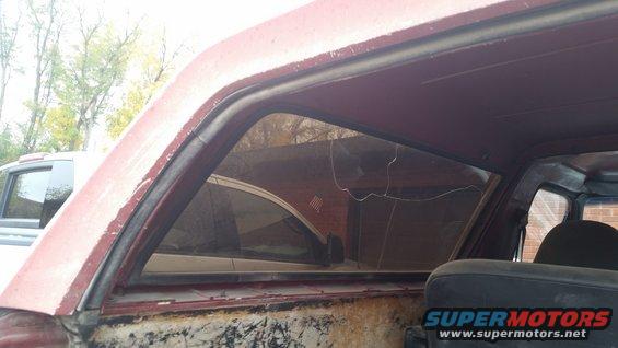 Close-up of a cracked rear quarter window on a red SUV, showing the seal and interior. Another SUV is visible through the window