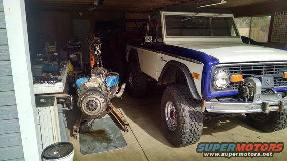 White and blue Ford Bronco with a winch on the front bumper, parked in a garage next to a detached engine