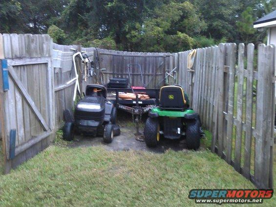 Two riding lawn mowers, one black and one green John Deere, parked next to a utility trailer in a fenced yard