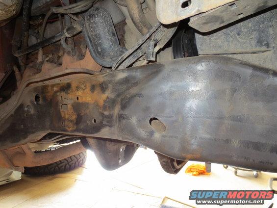 Underside view of a vehicle's frame and suspension parts, showing rust and wear