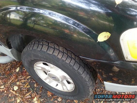 Front wheel and fender of a dark green Ford Explorer. Leaves are on the ground and on the vehicle