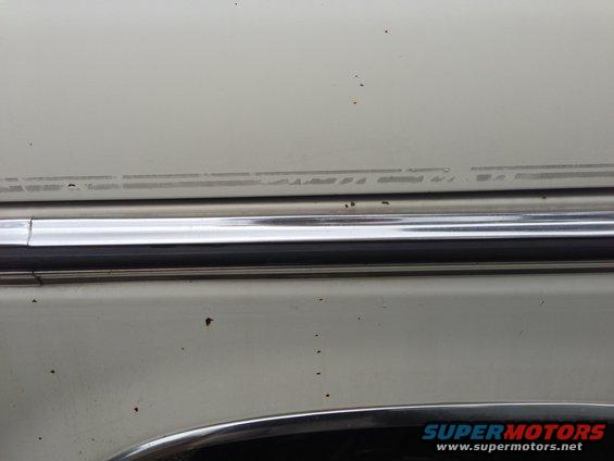 Close-up of light-colored car body with chrome trim and faint pinstripe detail. Watermark reads SUPERMOTORS