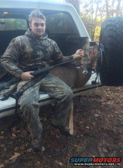 guy in camo sitting in back of truck with a deer and rifle