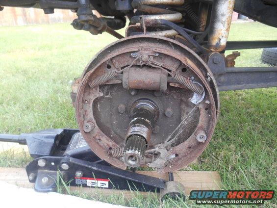 Rear axle brake drum assembly with drum removed, showing brake shoes and wheel cylinder. A floor jack is positioned underneath