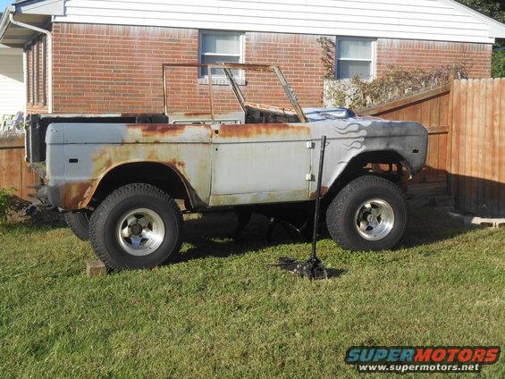 Old Ford Bronco body, rusty and missing parts, sitting in a grassy yard. Flames painted on the fender