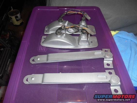 Two silver tail light assemblies with wiring and two silver metal brackets with pivot points, all on a purple plastic container