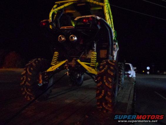 rear view of a yellow off-road buggy with large tires on a trailer, a white pickup truck is visible behind it at night