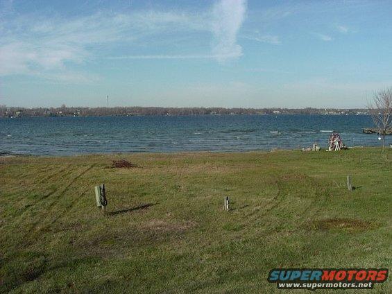 Grassy field with a body of water and a distant treeline under a cloudy sky
